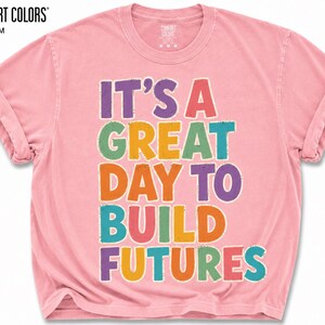 May include: A light pink Comfort Colors t-shirt with rolled-up sleeves. The shirt features the text "IT'S A GREAT DAY TO BUILD FUTURES" in colorful block letters. The brand name "COMFORT COLORS" and the color "BLOSSOM" are printed at the top.