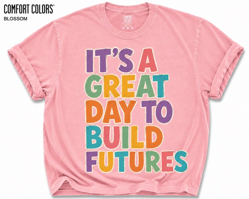 May include: A light pink Comfort Colors t-shirt with rolled-up sleeves. The shirt features the text "IT'S A GREAT DAY TO BUILD FUTURES" in colorful block letters. The brand name "COMFORT COLORS" and the color "BLOSSOM" are printed at the top.