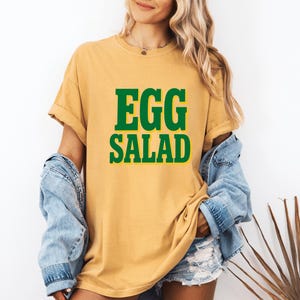 Peut inclure: T-shirt jaune moutarde avec les mots "EGG SALAD" en grandes lettres vertes et grasses avec un contour jaune. Le t-shirt est porté avec une veste en jean bleu clair et un short en jean. Le modèle sourit.