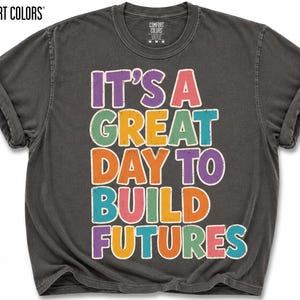 May include: A dark gray Comfort Colors t-shirt with rolled-up sleeves. The shirt features the text "IT'S A GREAT DAY TO BUILD FUTURES" in colorful, distressed lettering. The shirt is a medium size.