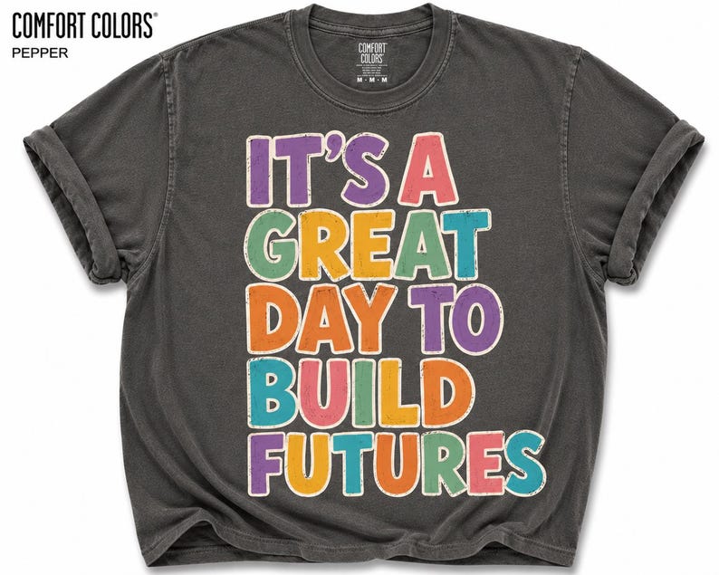 May include: A dark gray Comfort Colors t-shirt with rolled-up sleeves. The shirt features the text "IT'S A GREAT DAY TO BUILD FUTURES" in colorful, distressed lettering. The shirt is a medium size.