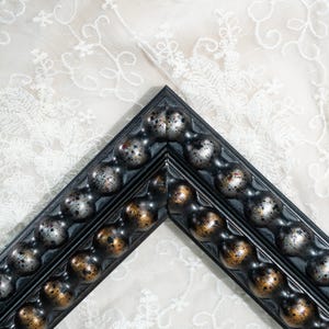 Può includere: Un angolo di due cornici sovrapposte con una modanatura a perline decorativa. Le cornici presentano una finitura scura con riflessi metallici argentati e bronzo anticato sulle perline arrotondate, creando un aspetto strutturato e variegato.