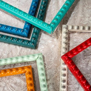 May include: A collection of rectangular picture frames featuring a beaded edge design. The frames are finished in various solid colours including bright turquoise, deep blue, dark teal, light mint green, vibrant orange, white, and glossy red.