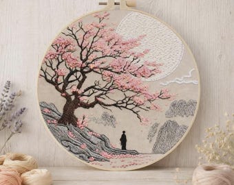 May include: Embroidered artwork in a round wooden hoop. The design features a cherry blossom tree with pink flowers, a full moon, and a figure standing on a rocky hillside. The color palette includes shades of pink, brown, gray, and cream.