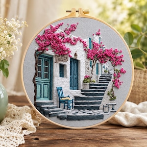 May include: A circular embroidery hoop featuring a detailed needlework scene of a white Mediterranean-style building with blue doors, a blue chair, and stone stairs, all adorned with vibrant pink bougainvillea flowers on a light gray fabric.