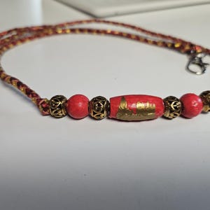 Puede incluir: Un cordón trenzado en rojo y dorado con un cierre de mosquetón plateado. El cordón está adornado con dos cuentas rojas redondas, dos cuentas rojas más pequeñas, cuatro espaciadores metálicos dorados ornamentados y una cuenta central roja en forma de barril.