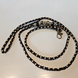 Puede incluir: Un cordón trenzado de color negro y beige, que presenta una serie de cuentas decorativas en tonos plateados, marrones y ámbar. El cordón incluye un cierre de mosquetón de metal en un extremo, proporcionando un punto de sujeción funcional.