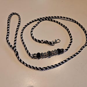 May include: A braided leash made of black and white cord, featuring a loop handle at one end and a metal swivel clasp at the other. The leash is accented with a series of decorative silver-toned metal beads and black glass beads.