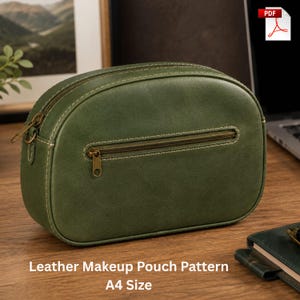 May include: An olive green leather makeup pouch featuring a rounded rectangular shape, a top zipper closure, and a horizontal zippered pocket on the front. The pouch is accented with visible light-colored stitching and antique brass-toned hardware.
