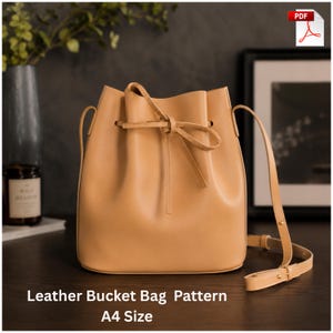 Puede incluir: Un bolso tipo cubo de cuero color canela con cierre de cordón y un lazo atado en la parte delantera, una correa larga ajustable para el hombro y un acabado liso. El texto "Leather Bucket Bag Pattern A4 Size" aparece en la parte inferior.