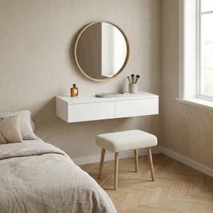 May include: A modern bedroom setup featuring a white floating vanity with two drawers, a round wooden-framed mirror, and a small upholstered stool. The room has a neutral color palette with a bed and a window.