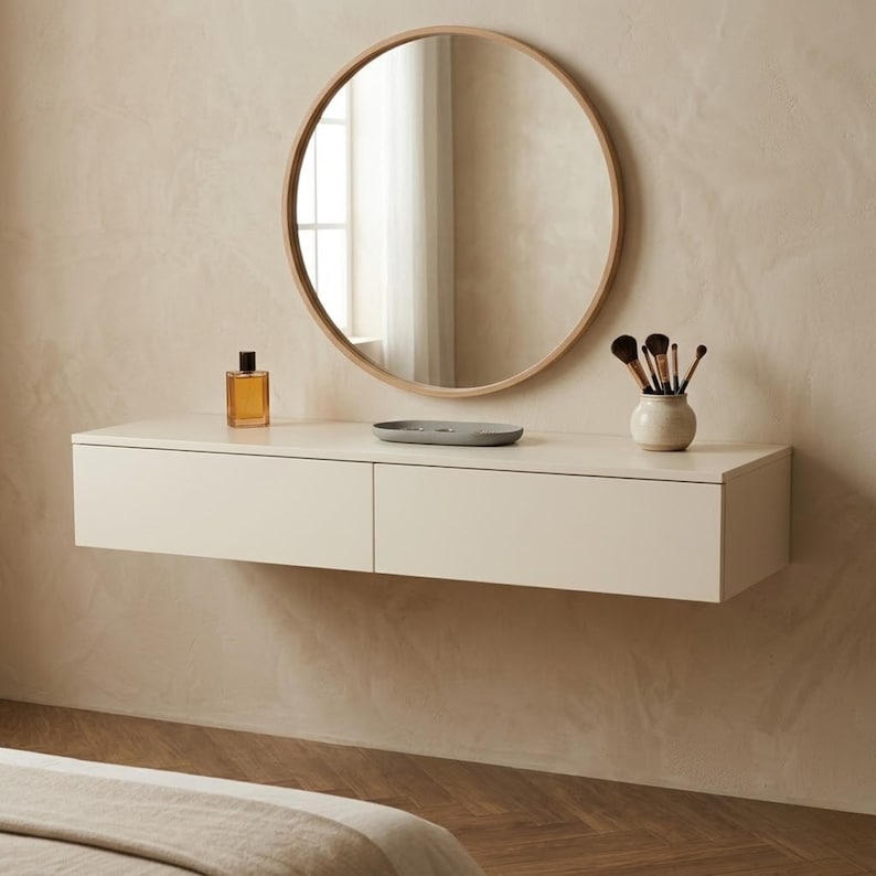 May include: A modern vanity setup featuring a round wooden-framed mirror above a floating cream-colored cabinet with two drawers. The cabinet holds a perfume bottle, a small gray dish, and a vase with makeup brushes. The wall is a neutral beige.