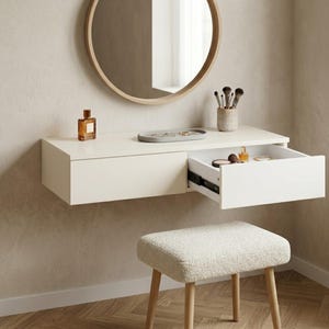 Puede incluir: Un tocador blanco flotante con un cajón abierto que revela cosméticos y joyas. Un espejo redondo cuelga sobre el tocador y un pequeño taburete con patas de madera clara se encuentra debajo. Un frasco de perfume y brochas de maquillaje están sobre el tocador.