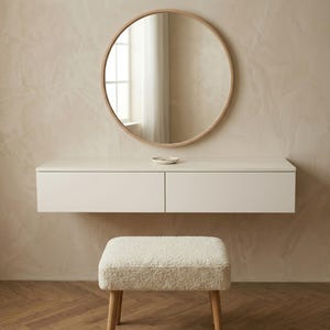 May include: A modern vanity set with a round mirror, a floating white cabinet with two drawers, and a matching upholstered stool. The mirror has a light wood frame. The stool has light wood legs and a cream-colored, textured seat.