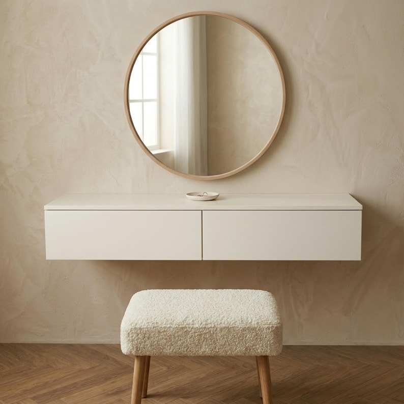 May include: A modern vanity set with a round mirror, a floating white cabinet with two drawers, and a matching upholstered stool. The mirror has a light wood frame. The stool has light wood legs and a cream-colored, textured seat.