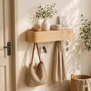 Könnte beinhalten: Helles Holz-Wandregal mit Haken, an dem eine beige Handtasche, Schlüssel, ein Schal und eine Sonnenbrille hängen. Eine Vase mit Grün, eine kleine Schale und eine dekorative Box befinden sich auf dem Regal. Ein Buch mit dem Titel "The Art of Home" ist ebenfalls vorhanden.