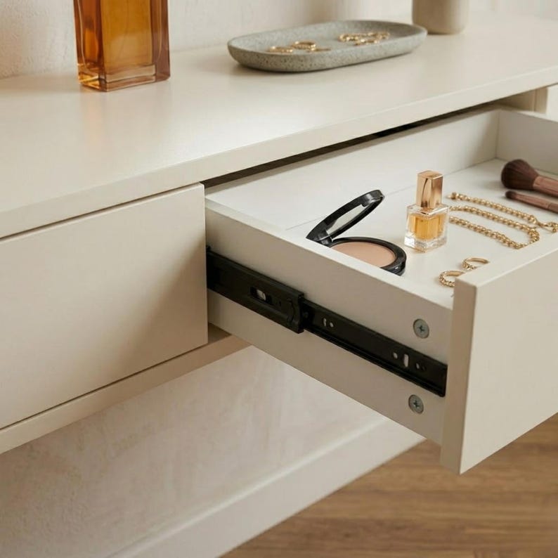 May include: A white vanity with an open drawer revealing makeup, perfume, and jewelry. A small perfume bottle and a compact are visible. A gold necklace and rings are also in the drawer. A tray with rings and a perfume bottle sit on top.