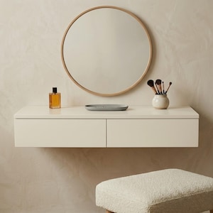 May include: A modern vanity set with a round mirror, a floating cream-colored drawer unit, and a small stool. The unit holds a perfume bottle, a gray tray, and a small vase with makeup brushes. The mirror has a light wood frame.