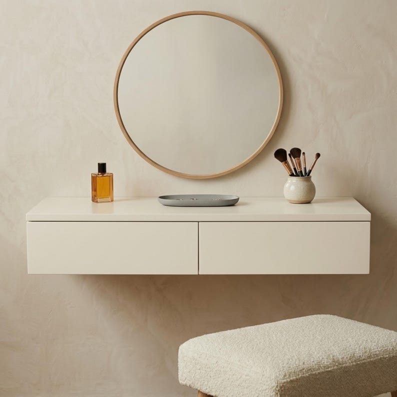May include: A modern vanity set with a round mirror, a floating cream-colored drawer unit, and a small stool. The unit holds a perfume bottle, a gray tray, and a small vase with makeup brushes. The mirror has a light wood frame.