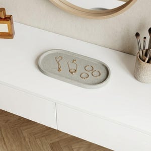 May include: A gray oval concrete tray holds gold jewelry, including a necklace, a ring, and hoop earrings. The tray sits on a white surface, with a perfume bottle and makeup brushes nearby. A wooden-framed mirror is in the background.