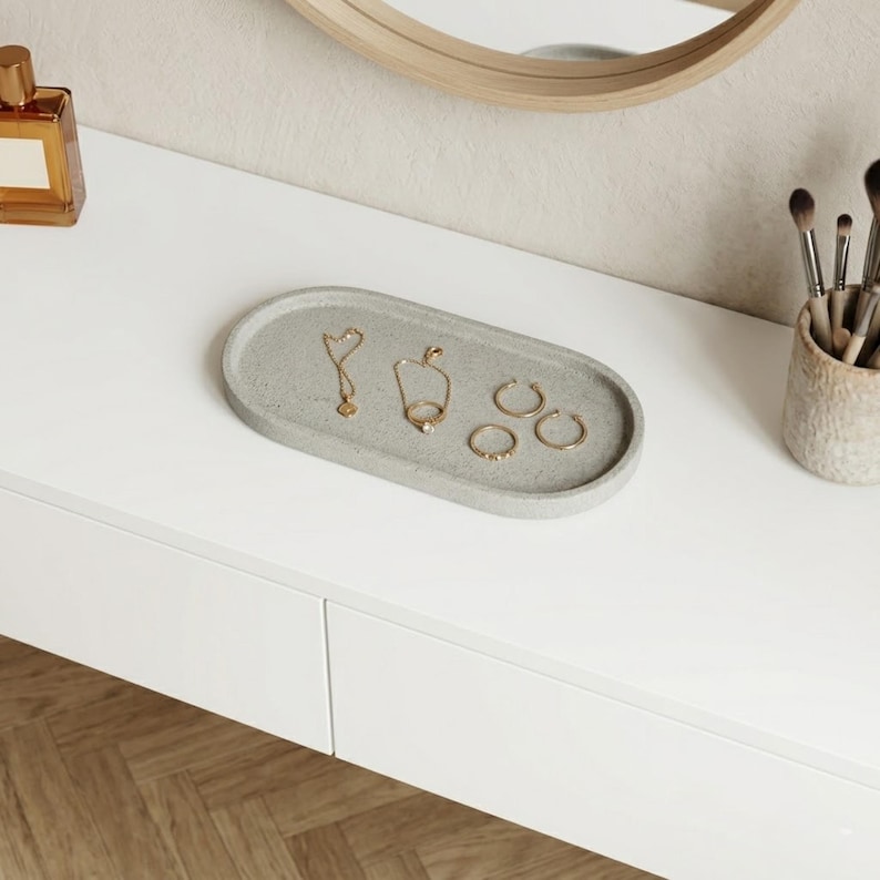 May include: A gray oval concrete tray holds gold jewelry, including a necklace, a ring, and hoop earrings. The tray sits on a white surface, with a perfume bottle and makeup brushes nearby. A wooden-framed mirror is in the background.