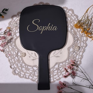 May include: A black hand mirror with the name "Sophia" in gold script. The mirror is on a white lace doily, with dried flowers and pink blossoms around it. The mirror has a rectangular shape with a handle.