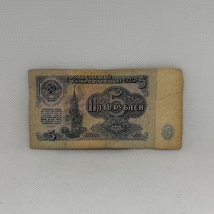 May include: A vintage 5 ruble banknote from the USSR, featuring a tan and black color scheme. The bill displays the Spasskaya Tower, the Soviet coat of arms, and the text "ГОСУДАРСТВЕННЫЙ КАЗНАЧЕЙСКИЙ БИЛЕТ СССР 5 ПЯТЬ РУБЛЕЙ 1961". It has a worn, textured appearance.