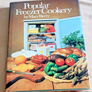 May include: A hardcover book titled Popular Freezer Cookery by Mary Berry. The cover features a photograph of an open freezer filled with food, alongside a wooden cutting board with an orange, sliced fruit, a knife, and fresh vegetables.