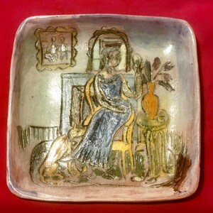 May include: A square ceramic dish featuring a relief illustration of a person seated on a chair next to a dog, a fireplace, and a small table with a vase of flowers. The scene is rendered in muted tones of blue, yellow, brown, and cream with a glossy finish.