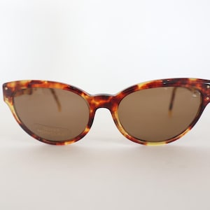 May include: Vintage tortoiseshell sunglasses with brown lenses. The cat-eye frames have a classic design with a speckled pattern in shades of brown and yellow. The lenses are a solid brown color, and the sunglasses have a small gold accent on each side.