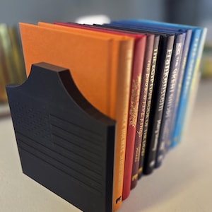May include: A black bookend featuring a raised American flag design on its front face, holding a row of hardcover books. The books include titles such as The Great Gatsby, Frankenstein, The Egyptian Book of the Dead, and The Book of Five Rings.