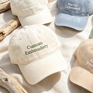 May include: Five baseball caps in cream, light blue, olive green, and tan. The cream cap features 'I complain about everything' in white, the blue cap 'Heavily Caffeinated' in white, the green cap 'Chill but...' in white, the tan cap 'Snack Provider' in white, and a second cream cap 'Custom Embroidery' in green.