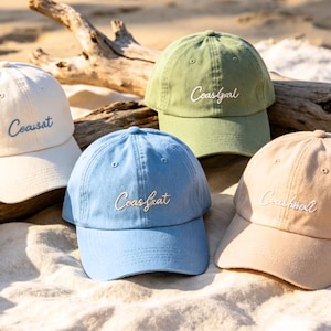 Könnte beinhalten: Fünf Baseballkappen in verschiedenen Farben: Weiß, Grün, Blau und Beige. Jede Kappe hat das Wort "Coasfgal" in Schreibschrift gestickt. Die Kappen sind auf einer sandigen Oberfläche mit einem Stück Treibholz angeordnet.