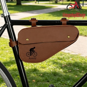 以下が含まれることがあります： フラップ開閉式の茶色の革製自転車フレームバッグ。ストラップで固定されています。バッグにはサイクリストのシルエットと「Avery」という名前がプリントされています。バッグは黒い自転車フレームに取り付けられています。