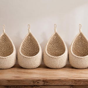 Puede incluir: Cuatro cestas colgantes beige en forma de lágrima hechas de material tejido. Cada cesta tiene un lazo en la parte superior para colgar. Están dispuestas en fila sobre una superficie de madera. Un jarrón con vegetación y una pequeña planta en maceta están a ambos lados.