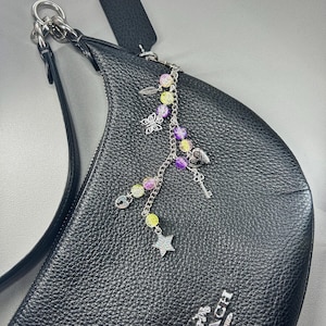 May include: Black leather handbag with a silver chain and charm. The charm features a butterfly, heart, key, and star. The handbag has a silver zipper and the brand name "COACH NEW YORK" is visible.
