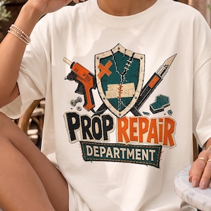 May include: An off-white t-shirt featuring a graphic print with a central shield, a glue gun, and a craft knife. The text reads "PROP REPAIR DEPARTMENT" in bold, distressed lettering. The design includes small hardware elements and a green foam block.