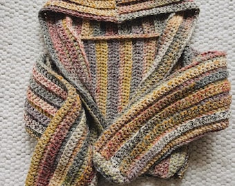 May include: A hooded scarf in a variety of colors, including yellow, pink, and gray. The scarf is handmade with a crochet stitch. The hood is folded over, and the scarf is spread out on a white, textured surface.