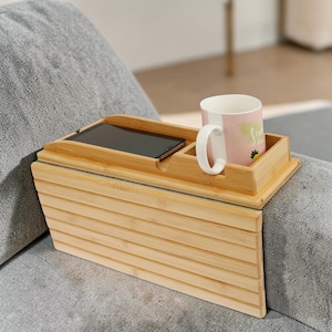 May include: A bamboo sofa arm tray with a built-in phone holder and a compartment for a mug. The tray is light brown and has a slatted design on the side. A pink and white mug with a handle sits in the compartment.