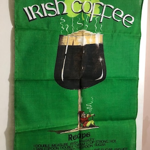 May include: A green tea towel with a recipe for Irish coffee. The recipe includes 1 double measure Irish whiskey, 1 tablespoon double cream, 1 cup strong hot black coffee, and 1 heaped teaspoon sugar. The towel has a shamrock design and a cartoon illustration of a person drinking Irish coffee.