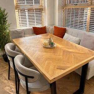 May include: A wooden dining table with a herringbone pattern, supported by black metal legs. Two grey upholstered chairs with black metal accents are positioned near the table. The table is surrounded by a built-in bench with orange and grey cushions. The room has large windows with wooden blinds.