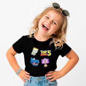 May include: Black t-shirt featuring colorful graphics from Toy Story 5. The design includes a retro handheld game, a blue hippo, a purple camera, and the Toy Story 5 logo in yellow and red.