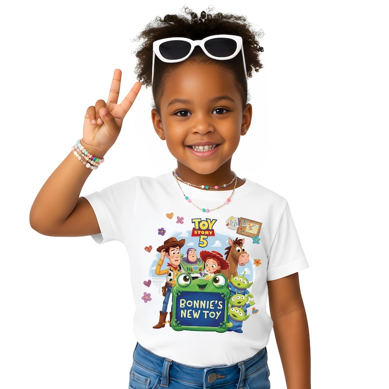 May include: White t-shirt featuring characters from Toy Story 5, including Woody, Buzz Lightyear, and Jessie. The shirt has the text "Bonnie's New Toy" on a green background. The child is wearing sunglasses and a necklace.