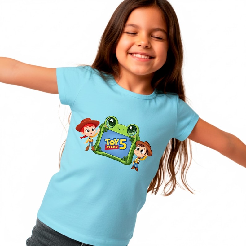 May include: A light blue t-shirt featuring a graphic of characters from Toy Story 5. The graphic includes Jessie, Woody, and a green frog-shaped frame with the movie title. The shirt is worn by a smiling child.
