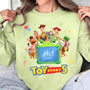 Pu&ograve; includere: Felpa verde chiaro con una grafica colorata di Toy Story, tra cui Woody, Buzz Lightyear e Jessie. Una cornice a forma di rana mostra il testo "Hi! Let's play!" con il logo "Toy Story 5" in basso.
