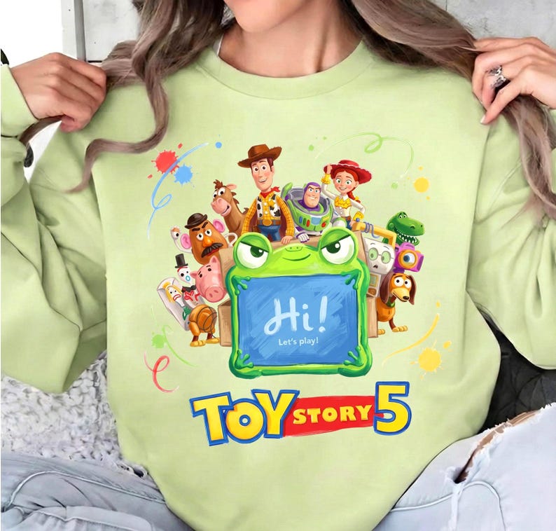 Pu&ograve; includere: Felpa verde chiaro con una grafica colorata di Toy Story, tra cui Woody, Buzz Lightyear e Jessie. Una cornice a forma di rana mostra il testo "Hi! Let's play!" con il logo "Toy Story 5" in basso.