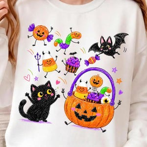 Op de afbeelding: Een wit sweatshirt met een kleurrijke grafische print in Halloween-thema. Het ontwerp bevat een zwarte kat, een pompoenemmer gevuld met snoep, een vleermuis, een candy corn-figuurtje, een cupcake en een worm, in een speelse, handgetekende stijl.
