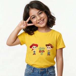 May include: A yellow t-shirt featuring the characters Jessie and Woody from Toy Story 5. The Disney Pixar logo is above the text "TOY STORY 5". The shirt is worn by a person wearing glasses and blue jeans.