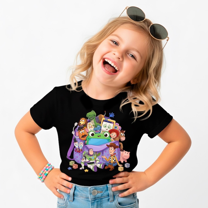 May include: Black t-shirt featuring a colorful graphic of various Toy Story characters emerging from a purple backpack. The design includes Woody, Buzz Lightyear, and other beloved characters. A fun and playful design for kids.