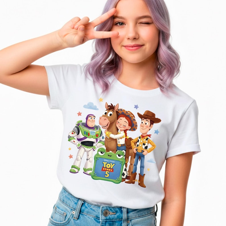 May include: White t-shirt featuring characters from the Toy Story franchise, including Buzz Lightyear, Woody, Jessie, and a horse. The shirt has the text "Toy Story 5" on a green frog-shaped sign. The design is surrounded by stars and clouds.